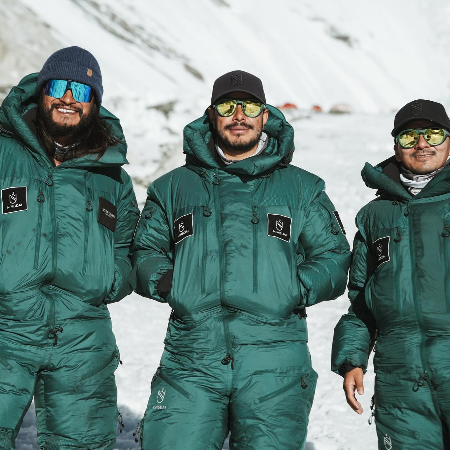 Three men including nims purja wearing matching nimsdai store dark green technical snow suits and winter gear, including reflective sunglasses and black hats, standing in a snowy mountain setting.tore